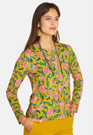 Woman wearing a mustard yellow floral top with pink flowers, matching mustard pants, gold earrings, and long layered necklaces.