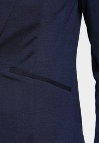 Blazer bleu marine avec une texture de tissu lisse, un design structuré, et une poche latérale avec un détail de bordure élégante.