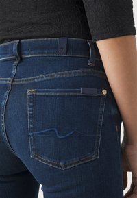 Jeans de denim azul oscuro con una cintura de altura media, que presentan un diseño de bolsillo trasero y detalles de costura azul. La terminación incluye una etiqueta de marca.