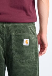 Green corduroy pants with a back pocket featuring a white label with an orange logo. Texture is ridged; design includes a straight cut.