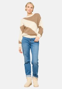 Pleteno pulover v kremni in rjavi barvi z diagonalnimi črtami, v kombinaciji z svetlo modrimi kavbojkami in pevskimi čevlji iz ovčje kože. Mehak material in sproščen kroj.