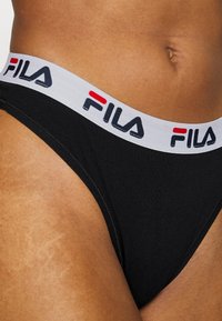 Crne pamučne gaće s bijelim elastičnim pojasom na kojem se nalazi "FILA" logo u crvenoj i crnoj boji. Glatka tekstura, minimalistički dizajn.