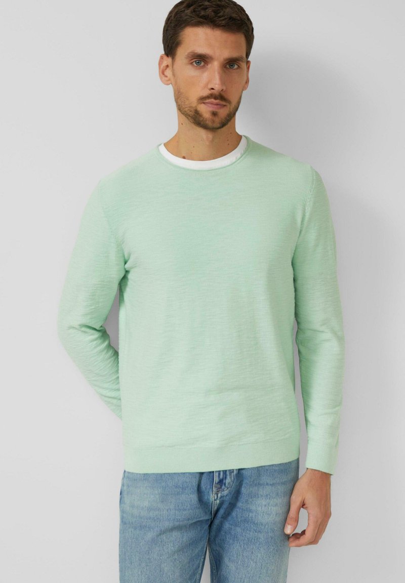 Man draagt een lichtgroene trui met lange mouwen over een wit overhemd en blauwe spijkerbroek, staande tegen een effen lichtgrijze achtergrond.