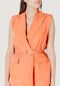 Blazer senza maniche arancione con scollo a V profondo, vita cinturata, due tasche anteriori e finitura testurizzata. Dettagli metallici vicino alle tasche.