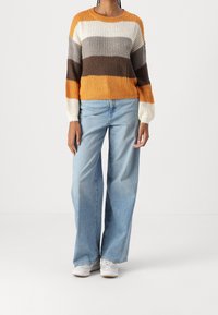 Person, der einen Strickpullover mit horizontalen Streifen in Orange, Weiß, Grau und Braun trägt, kombiniert mit hellblauen, weit geschnittenen Jeans und weißen Sneakers.