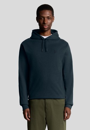 Dark blue cotton hoodie with drawstrings, ribbed cuffs, and a small embroidered logo on the chest. Worn with olive green pants.