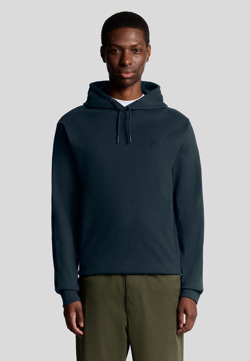 Felpa in cotone blu scuro con lacci, polsini a costine e un piccolo logo ricamato sul petto. Indossata con pantaloni verde oliva.