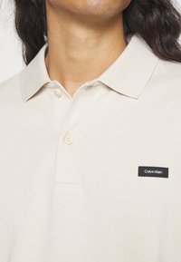 Homme portant un polo beige Calvin Klein avec deux boutons et une étiquette logo noire sur la poitrine gauche.