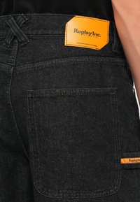 Black denim jeans featuring a distinctive orange label with "Replay Inc." on the waistband. The fabric has a soft, textured look.