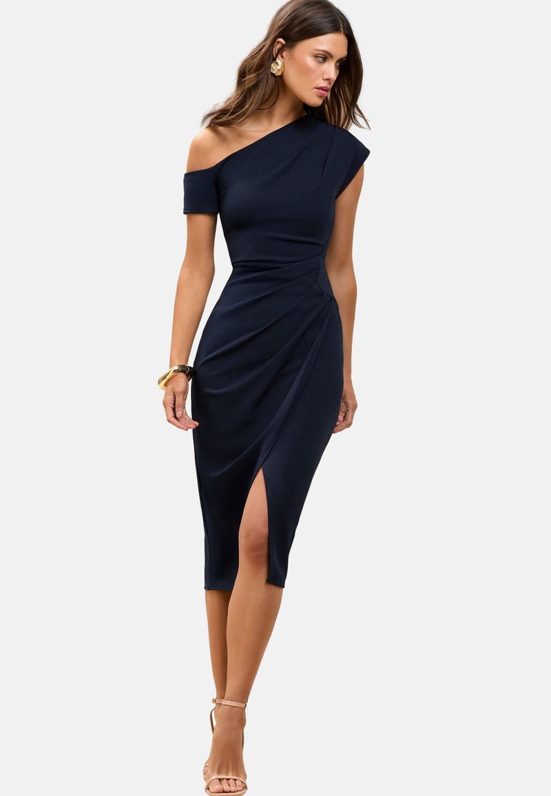 Frau trägt ein marineblaues One-Shoulder-Midikleid mit seitlichem hohem Beinschlitz, goldene Creolen und nudefarbene Absatzsandalen mit Knöchelriemen.