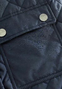 Gestepptes dunkelblaues Jacket mit Wassertropfen auf einer glatten Stoffoberfläche, ausgestattet mit einer rechteckigen Tasche und silbernen Druckknöpfen.