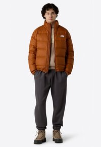 Polstret brun jakke med høj krave og lynlås, båret over en beige sweatshirt, kombineret med grå joggingbukser og tan-farvede støvler.