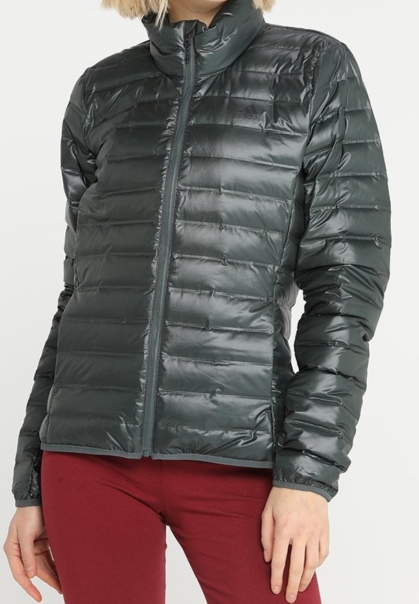 Žena v lesklé tmavě šedé prošívané bundě se zipem vpředu a vysokým límcem, v kombinaci s těsnými tmavě červenými kalhotami.