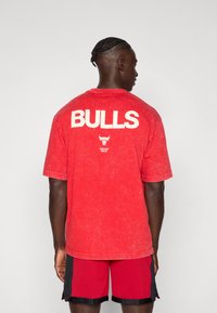 Mand med fletninger iført en rød Chicago Bulls T-shirt og røde shorts, vendende væk mod en ensfarvet lys baggrund.