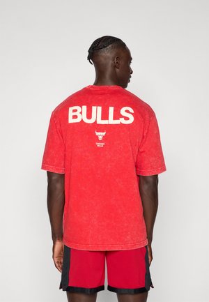 Mand med fletninger iført en rød Chicago Bulls T-shirt og røde shorts, vendende væk mod en ensfarvet lys baggrund.
