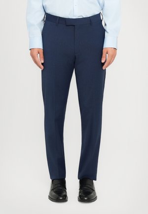 Man wearing navy blue formal trousers, light blue dress shirt, and black leather loafers, standing against a plain light background.