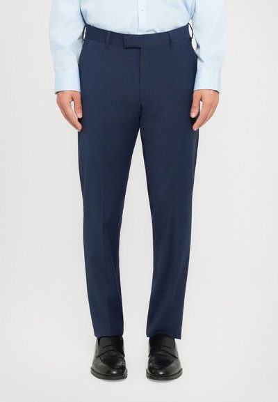 Mann in navyblauen formellen Hosenträgern, hellblauem Hemd und schwarzen Leder-Lofers, der vor einem schlicht hellen Hintergrund steht.