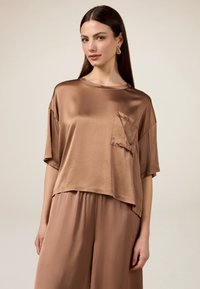 Top corto in satin marrone con maniche corte, scollo rotondo e taschino a sinistra, abbinato a pantaloni larghi coordinati.
