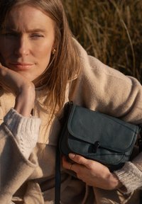 Borsa a tracolla in pelle color turchese con chiusura a pattina, dettaglio della striscia frontale e cuciture a contrasto, tenuta contro un maglione beige chiaro.