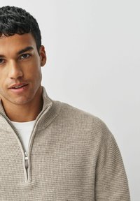 Beige Strickjacke mit Reißverschluss und Texturmuster sowie hohem Kragen, getragen über einem weißen Hemd. Nahaufnahme des Oberkörpers und des Gesichts.