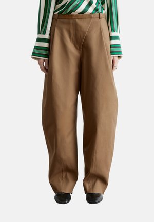 Person trägt breite braune Hose, grün-weiß gestreiftes langärmeliges Hemd und schwarze Schuhe, steht vor einem weißen Hintergrund.