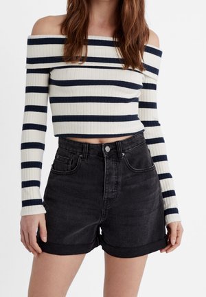Kvinde iført en hvid off-shoulder ribstrikket sweater med sorte vandrette striber og sorte denimshorts med oprullede kanter.