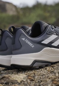 Chaussures de trail running Terrex grises et noires avec semelles intermédiaires blanches sur un terrain extérieur rocheux.