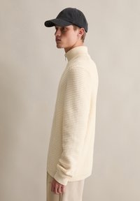 Cremfarbener Strickpullover mit hohem Kragen und gerippten Bündchen, kombiniert mit einer dunklen Baseballkappe. Glatte Textur mit Waffelmuster.