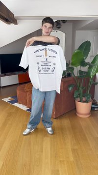 Bela bombažna majica s kratkimi rokavi in grafiko "VINTAGE VIBES" v modri barvi, z papirnato etiketo. Model nosi modre kavbojke in srebrne čevlje.