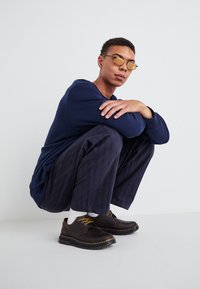 Brown leather shoes with yellow stitching, navy long-sleeve shirt, and striped dark pants. Subject is crouching against a light background.