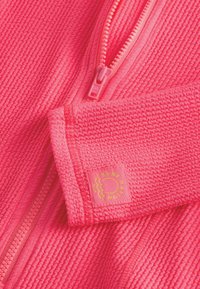 Bright pink zip-up jacket with textured surface, featuring a matching zipper and a square tag with yellow sun logo on the sleeve.
