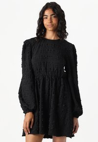 Vestido negro de textura con un cuerpo fruncido, mangas largas abullonadas y una silueta fluida. La tela presenta un diseño tejido y estampado.