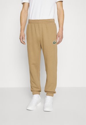 Homme portant un pantalon de survêtement beige avec des poignets élastiques et un logo crocodile vert, des baskets blanches et un t-shirt blanc, debout sur un fond uni.