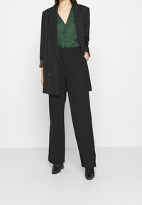 Blazer nero oversize, camicia verde in satin con bottoni, e pantaloni neri larghi. Stivali con tacco neri. Texture lisce, vestibilità su misura.