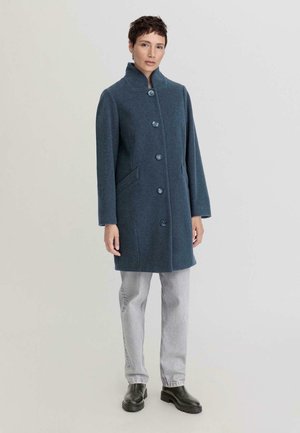 Cappotto di lana blu con colletto alto, bottoni frontalmente e tasche laterali, abbinato a jeans grigi e stivaletti neri. Texture liscia, design minimal.