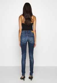 Jeans ajustados de denim azul oscuro, con una cintura de altura media, cinco bolsillos y una sutil textura tejida. Top negro de tirantes debajo.