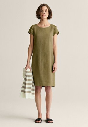 Nainen, jolla on lyhyet ruskeat hiukset, pukeutunut oliivinvihreään polvipituiseen mekkoon ja mustiin sandaaleihin, pitää vasemmassa kädessään kerma-beige raidallista neulepaitaa.