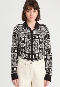 Chemise noire à boutons avec un motif floral et géométrique blanc, un col noir et des manches longues. Portée avec un pantalon de couleur claire.