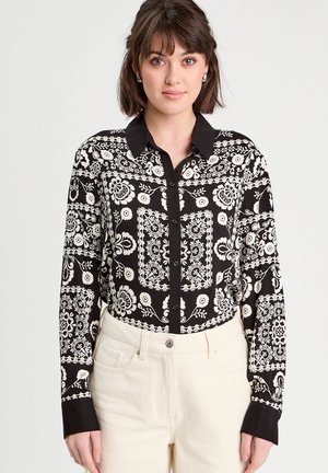 Chemise noire à boutons avec un motif floral et géométrique blanc, un col noir et des manches longues. Portée avec un pantalon de couleur claire.