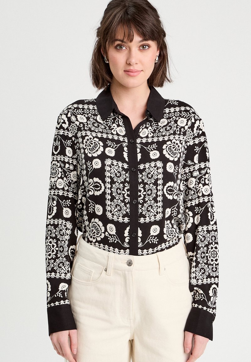 Chemise noire à boutons avec un motif floral et géométrique blanc, un col noir et des manches longues. Portée avec un pantalon de couleur claire.