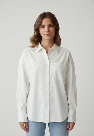 Mujer con cabello castaño ondulado, vistiendo una camisa blanca suelta abotonada y jeans azul claro, posando frente a un fondo liso de color gris claro.