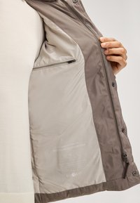 Grijze geïsoleerde jas met een tan voering, voorzien van een ritszak, drukknoopsluitingen en een label dat aangeeft dat hij gevuld is met 100% gerecycled polyester.