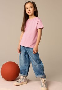 Maglietta rosa chiaro a maniche corte, pantaloni larghi in denim blu e sneaker alte con motivo. Una grande palla arancione si trova vicino, su uno sfondo chiaro.