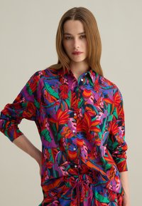 Camisa de botón colorida con un atrevido patrón floral en rojo, verde y púrpura. La tela tiene una textura suave y un corte relajado.