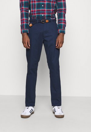 Man wearing navy blue pants, a woven navy belt with tan leather details, white sneakers with black stripes, and a red and green plaid shirt.