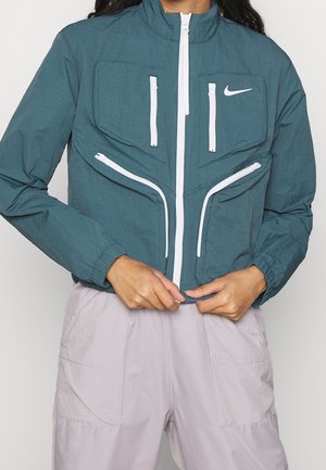 Personne portant une veste Nike turquoise avec quatre poches verticales blanches zippées et un pantalon gris clair, mains tenant le devant de la veste.