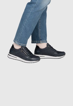 Persoon draagt zwarte sneakers met blauw patroon en witte zolen, lichtroze sokken en opgerolde blauwe spijkerbroek, tegen een effen lichte achtergrond.