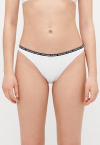 Hvit bikini-bunn med svart elastisk midjebånd som har teksten "Copenhagen Studios." Glatt stoff, design med minimal dekning.