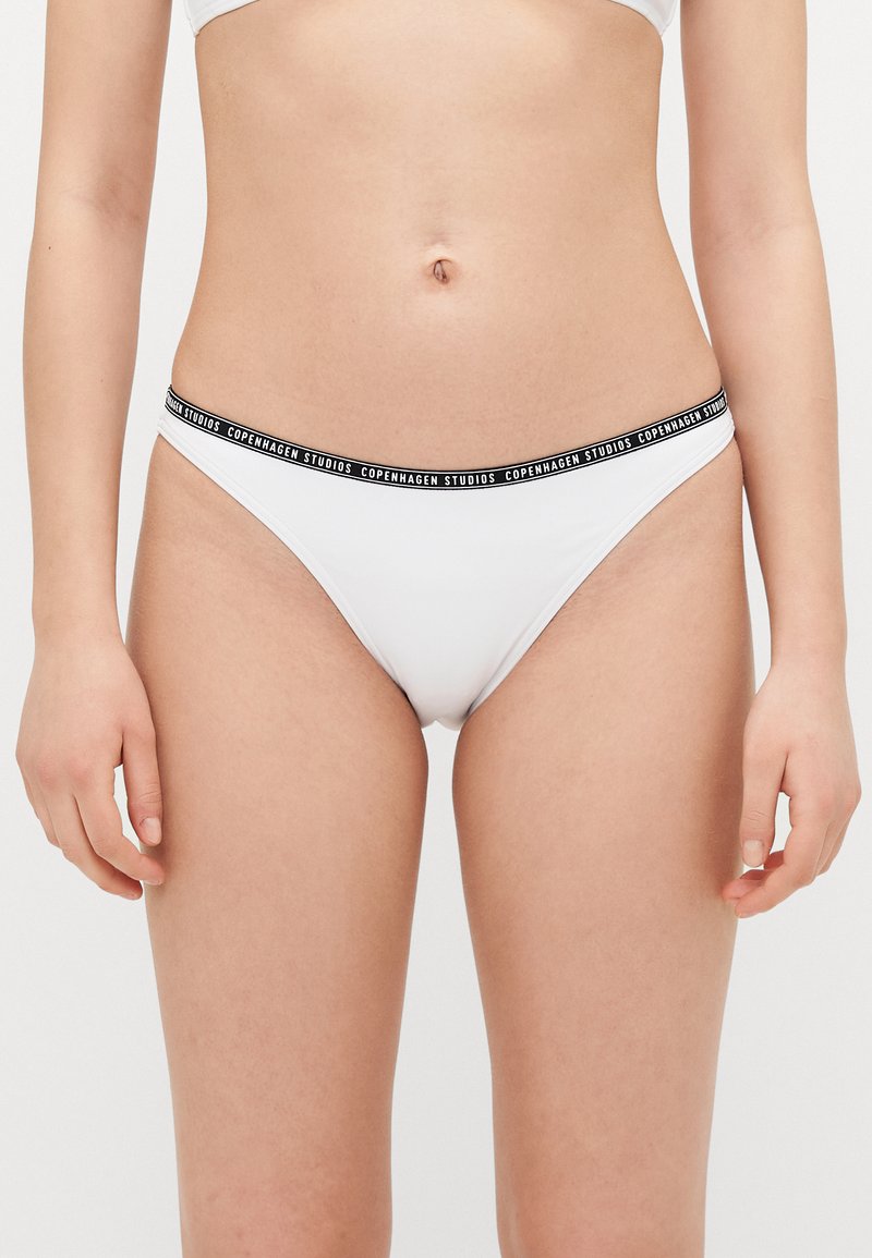 Hvit bikini-bunn med svart elastisk midjebånd som har teksten "Copenhagen Studios." Glatt stoff, design med minimal dekning.