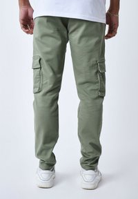 Pantalones cargo verde claro con bolsillos laterales, hechos de una tela suave. Diseño de pierna ajustada, combinados con zapatillas blancas para contraste.